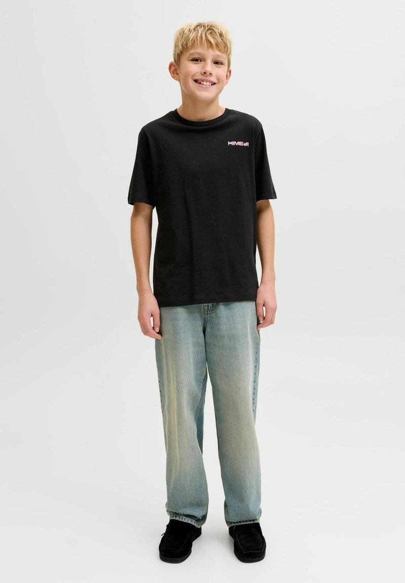 Glunderende jongen staand, dragend een zwart grafisch T-shirt, lichtblauwe spijkerbroek en zwarte schoenen tegen een effen witte achtergrond.