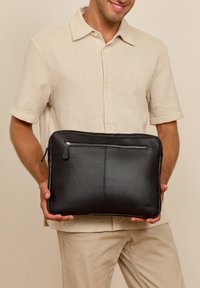 Vīrietis bēšā kreklā un biksēs, tur melnu ādas klēpjdatora futrāli ar priekšpuses rāvējslēdzēja kabatu pie vidukļa līmeņa.