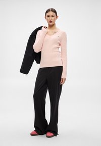 Rozā kabeļu raksta džemperis ar V-veida kaklu, melnas svītrains bikses un rozā slip-on apavi. Modelis tur melnu jaku pār plecu.