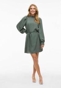 Groene, gestructureerde jurk met lange, opgeblazen mouwen en een strik rond de taille. Boven de knie lengte, met een hoge halslijn en een gladde stof.