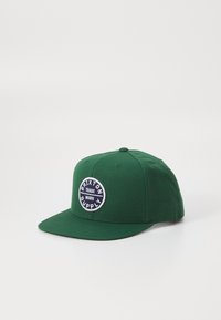 Green cotton baseball cap with a flat brim, featuring a round embroidered patch labeled "Brixton Supply" in white and navy.