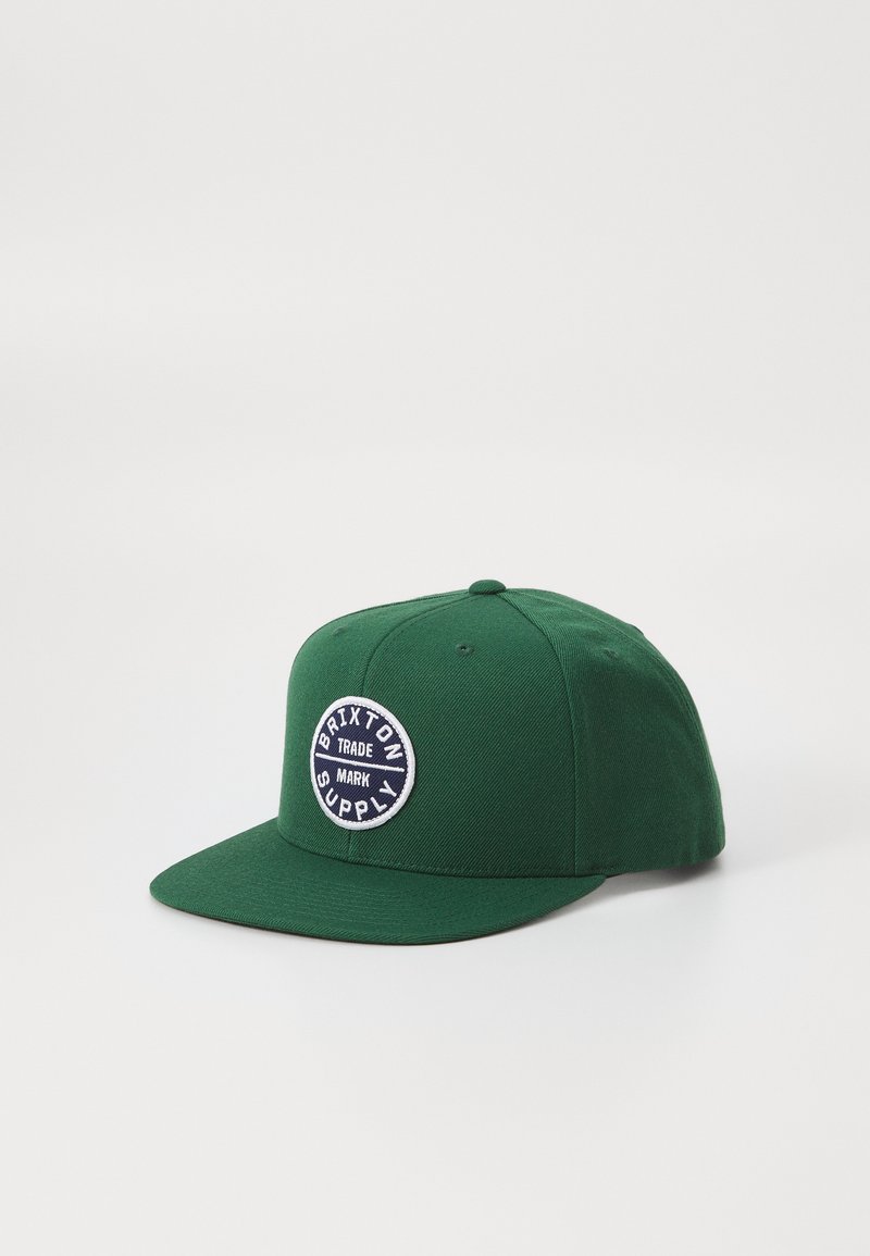 Green cotton baseball cap with a flat brim, featuring a round embroidered patch labeled "Brixton Supply" in white and navy.
