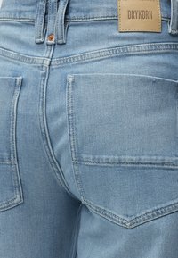 Jean en denim bleu clair vu de dos avec coutures visibles, deux poches arrière, passants pour ceinture, rivet en cuivre et une étiquette de marque beige portant la mention "DRYKORN."