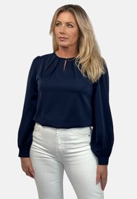 Granatowa bluzka z długimi, bufiastymi rękawami i dekoltem w kształcie łezki. Noszona z białymi dżinsami, tworząc kontrast kolorystyczny.