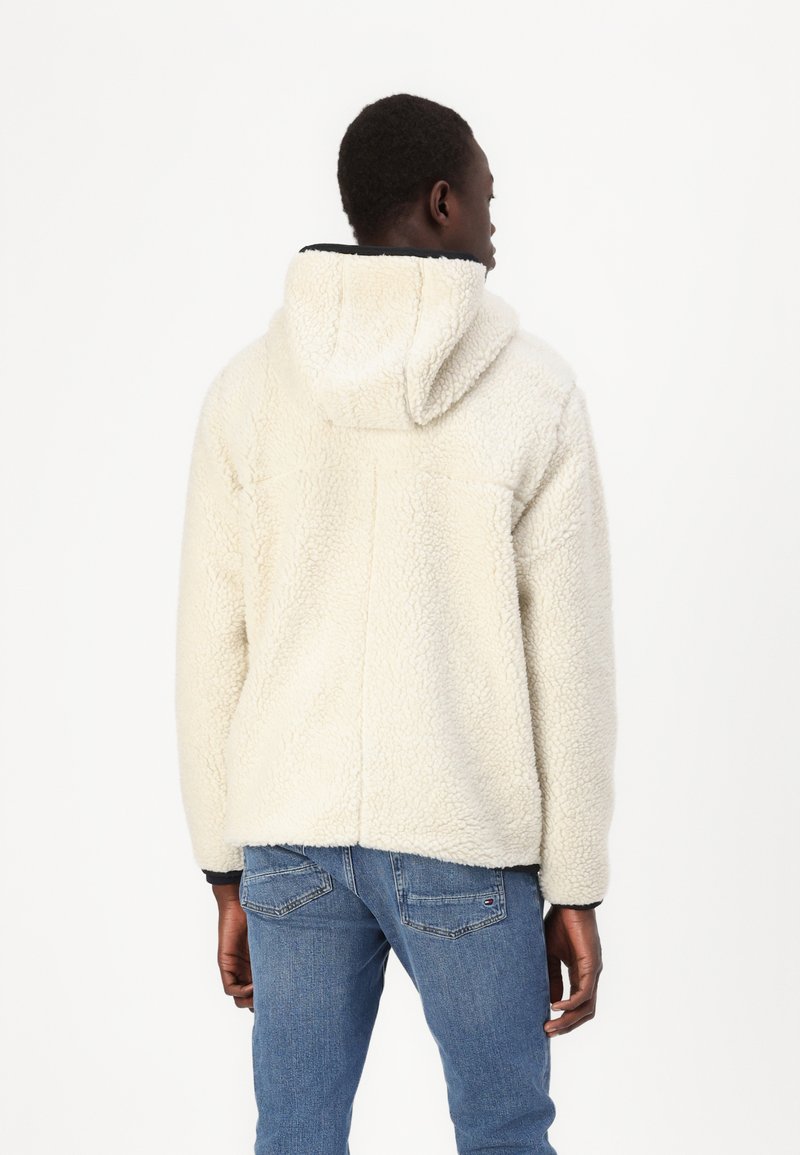 Veste en polaire couleur crème avec capuche, surface texturée et coupe décontractée. Détails de couture visibles et ourlet inférieur.