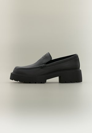 Mocassin en cuir noir avec semelle épaisse, talon bloc et petit logo métallique sur la tige, présenté de profil sur un fond neutre.