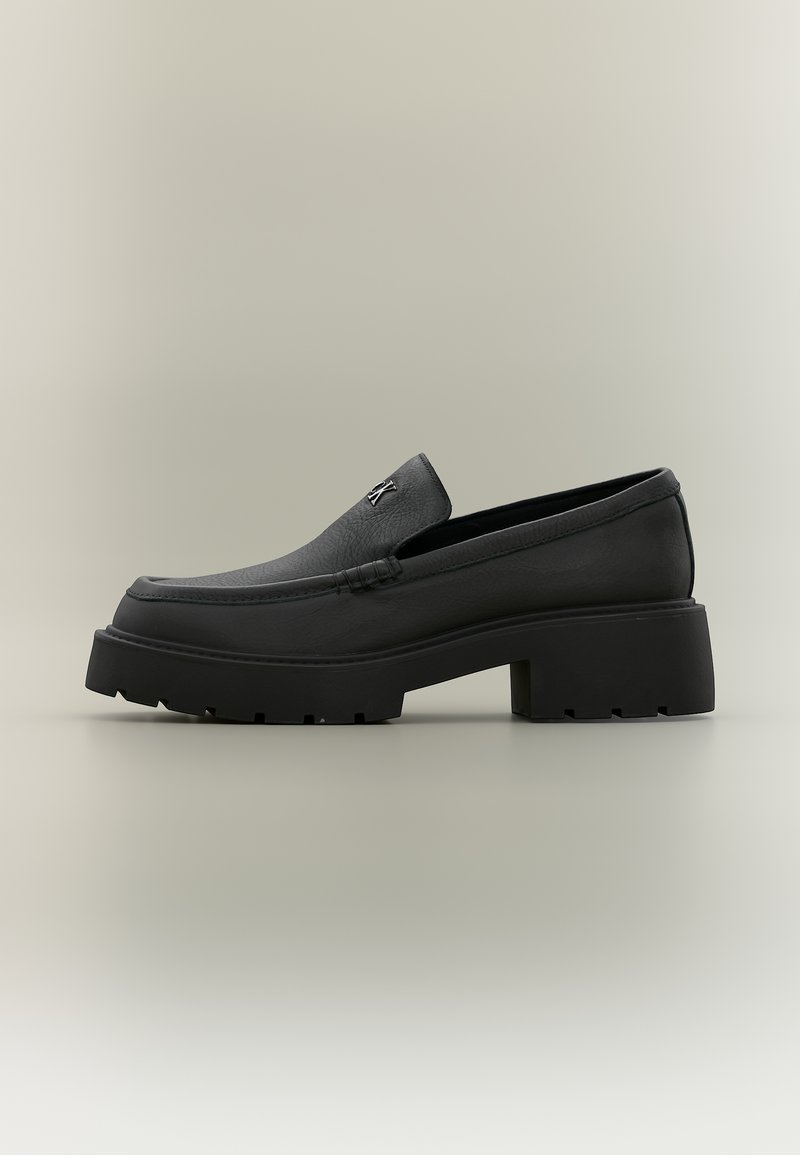 Mocassin en cuir noir avec semelle épaisse, talon bloc et petit logo métallique sur la tige, présenté de profil sur un fond neutre.
