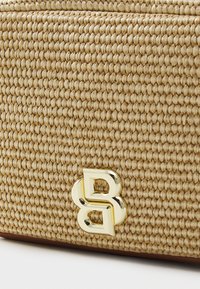 Tkaná slaměná kabelka v zlatavě béžové barvě s texturovaným povrchem. Disponuje výrazným zlatým logem na přední straně.