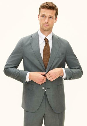 Man in grijs pak, wit overhemd en bruin patroon stropdas, sluit een knoop van zijn jasje terwijl hij iets schuin naar de zijkant kijkt tegen een effen achtergrond.