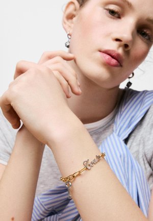 Persona joven con piel clara que lleva una pulsera de cadena dorada con una llave y la palabra "Confianza", vestida con una camisa gris y tela a rayas azules.