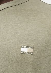 Λαδί βαμβακερό t-shirt με στρογγυλή λαιμόκοψη, που διαθέτει ένα μικρό ορθογώνιο ένθετο με την επιγραφή "TOMMY JEANS" σε λευκά και μπεζ γράμματα.