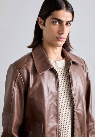 Giacca di pelle marrone con colletto ampio, chiusura con zip e tasche laterali. Indossata sopra un maglione lavorato a maglia beige chiaro con un motivo testurizzato.