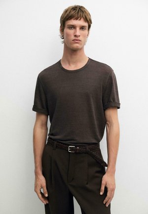 Junger Mann trägt ein dunkelbraunes T-Shirt mit hochgekrempelten Ärmeln, das in hoch taillierte dunkle Hose mit geflochtenem Gürtel gesteckt ist, vor schlichtem hellem Hintergrund.