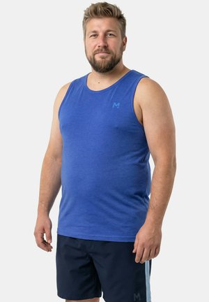 Bearded man wearing a blue sleeveless shirt and dark shorts standing against a plain light background, facing forward.