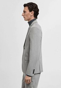 Giacca da uomo grigia chiara con un design strutturato, monopetto, con revers a notch e bottoni neri, indossata su un dolcevita scuro.