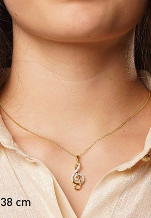 Goldene Halskette mit einem Notenschlüssel-Anhänger, geschmückt mit kleinen, klaren Kristallen, um den Hals einer Person getragen, die ein hell gefärbtes, strukturiertes Hemd trägt.