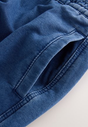 Mörkblå denimjoggers med mjuk textur, utrustade med elastisk midjeband och sidofickor. Sömmen är synlig längs kanterna.
