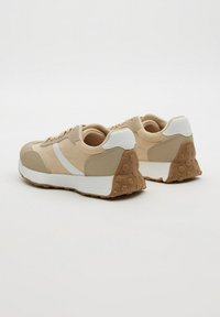 Zapatillas bajas beige y blancas con suelas de goma que presentan un patrón de almohadillas hexagonales, vistas desde la parte trasera sobre un fondo blanco liso.