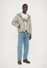 Helehall triibuline jope, tõmblukuga, mustade detailidega, kantud üle valge graafilise t-särgiga, kombineeritud helesiniste teksade ja pruuni spordijalatsitega.