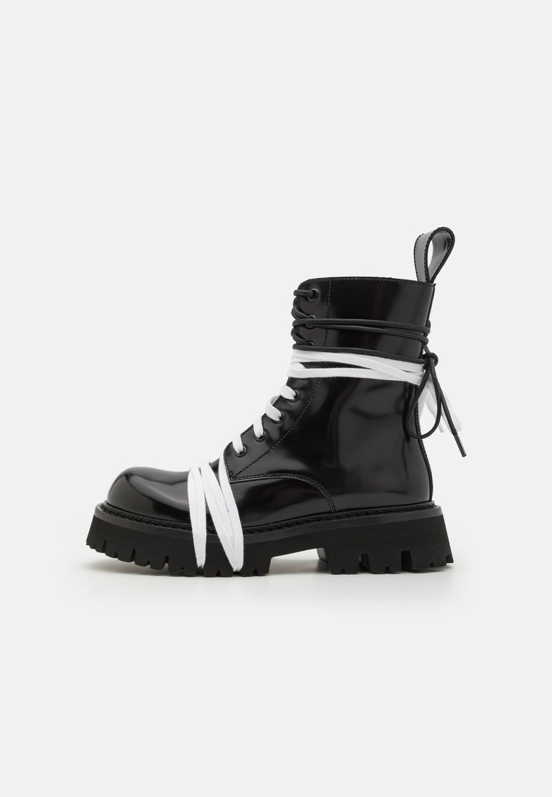 Bota de charol negra con suela de goma gruesa, cordones blancos enrollados, ojetes y una lengüeta en la parte posterior.