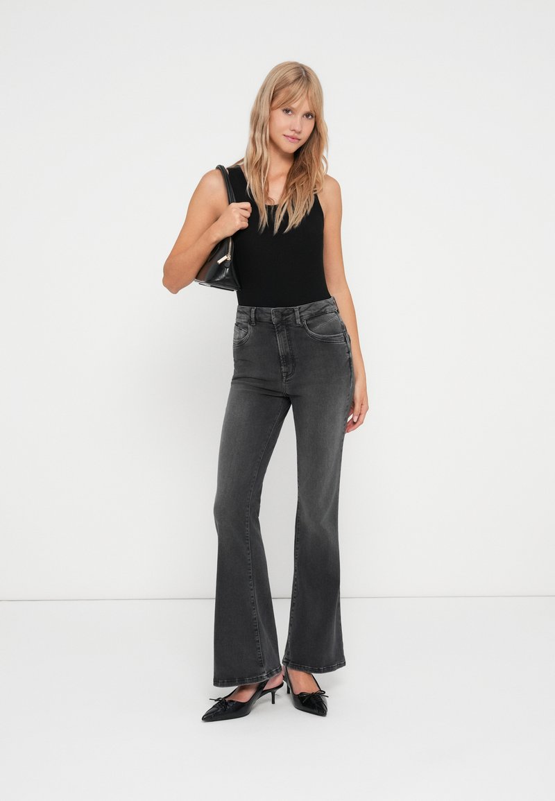 Schwarz hochtaillierte, ausgestellte Jeans mit frontalen Taschen und Dufthier wie verwaschenem Stoff, kombiniert mit einem figurbetonten schwarzen Tanktop und schwarzen spitzen High Heels.