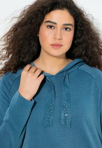 Donna con lunghi capelli ricci indossa una felpa teal con lacci decorati con strass, mentre tiene un laccio vicino alla spalla.