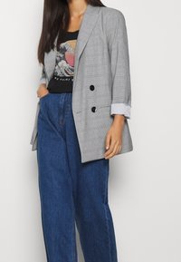 Grå rutig dubbelknäppt kavaj med svarta knappar, uppvikt ärmar, parat med en grafisk svart t-shirt och högmidjade blåjeans.