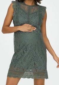 Grünes Spitzen-Maternity-Kleid mit gewellten Ärmeln, floralen Mustern und einem strukturierten Oberteil, ergänzt durch einen fransigen Spitzen-Saum.