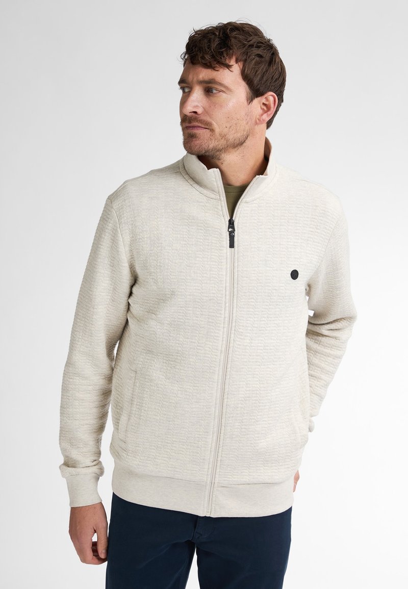 Jasno beżowa kurtka na zamek błyskawiczny o teksturowanej powierzchni, z wysokim kołnierzem, bocznymi kieszeniami oraz małym czarnym logo na piersi. Wykonana z miękkiego materiału.