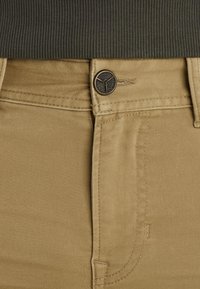 Tan pants made of smooth fabric featuring a zipper fly and a decorative button. Includes side and back pockets, with subtle stitch detailing.