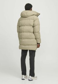 Long, puffy down parka in light olive green with a hood. Features vertical quilting and a zip closure, paired with dark cargo pants and sneakers.