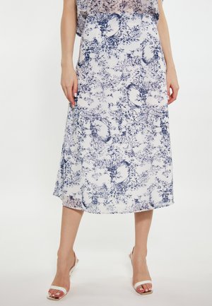 Donna che indossa una gonna midi bianca con motivo floreale e di uccelli blu, abbinata a tacchi aperti bianchi, in piedi davanti a uno sfondo uniforme.