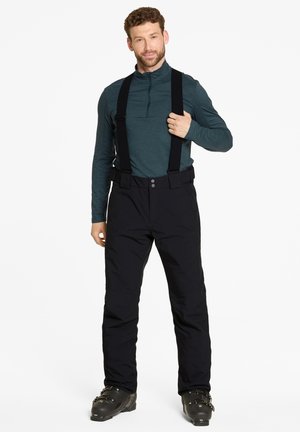 Black snow pants with adjustable suspenders, two front buttons, and a loose fit. Worn over a dark teal long-sleeve shirt and black ski boots.