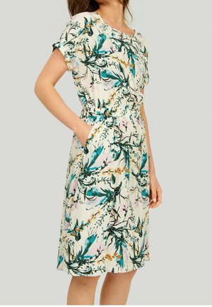 Vrouw draagt een witte jurk tot op de knie met een groen, blauw en geel bloemenpatroon, korte mouwen en een elastische taille, handen in de zakken.
