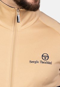 Beige zip-jakke med høj krave, med et navy broderet logo på brystet og et glat, struktureret stof.