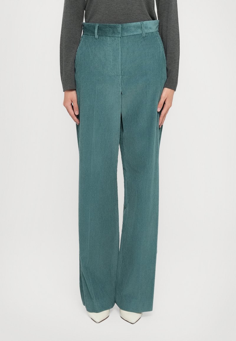 Wide-leg corduroy trousers in teal, featuring prominent vertical stripes, a belted waist, and a soft texture. Styled with white pointed heels.