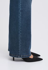 Jean en denim bleu foncé à coupe droite, ourlet retroussé, associé à des escarpins en suède noir à bout pointu sur un fond clair.