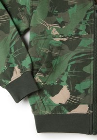 Giacca mimetica verde con zip frontale, polsini a coste, tasca con chiusura a bottone e tessuto testurizzato che presenta varie sfumature di verde e beige.