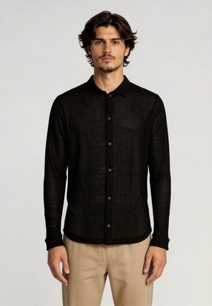 Homme aux cheveux mi-longs portant une chemise noire transparente boutonnée et un pantalon beige, debout devant un fond clair uni.