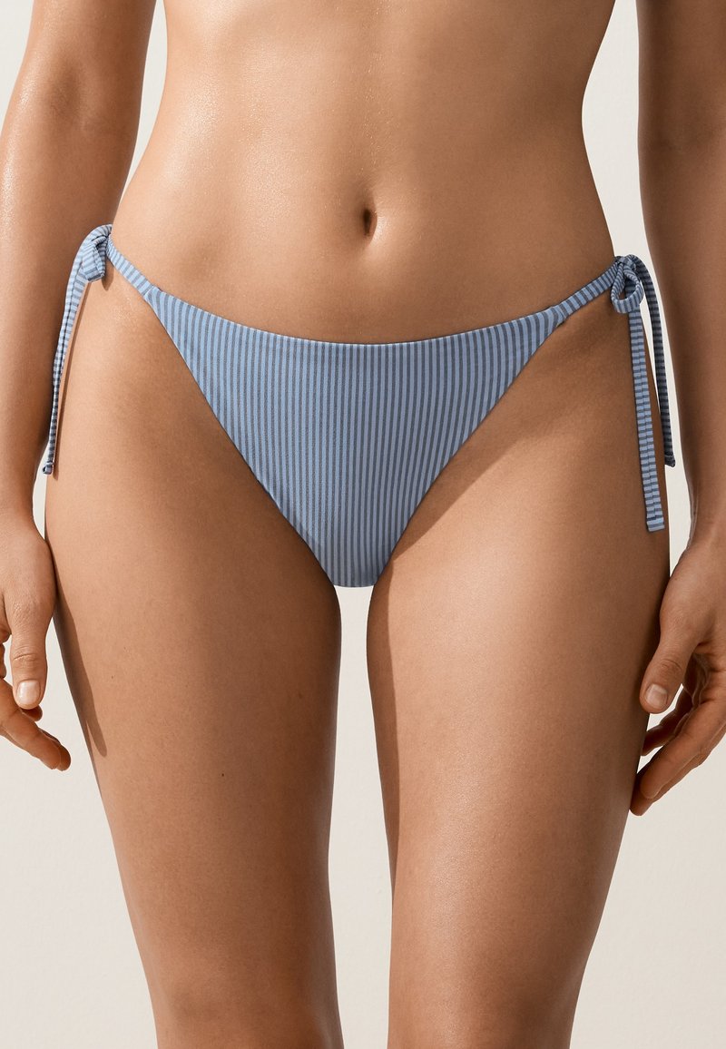 Femme portant un bas de bikini rayé bleu et blanc avec des détails noués sur les côtés, montrée de la taille jusqu'au milieu de la cuisse.