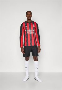 Schwarz-rotes langärmeliges Fußballtrikot mit gestreiftem Design, versehen mit einem Logo und Text, kombiniert mit schwarzen Shorts und weißen Socken.