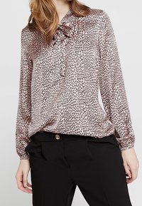 Blouse en imprimé léopard en tissu léger avec un nœud au col. Manches longues avec des poignets froncés. Associée à un pantalon noir.