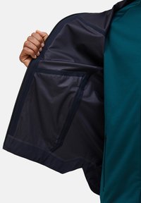 Mano che tiene aperta una giacca blu navy per mostrare una grande tasca interna in rete con una camicia turchese visibile sotto.