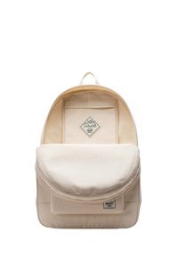 Beige canvas backpack with a rounded top, front pocket, and a flat flap. Features a logo tag and a top handle for carrying.