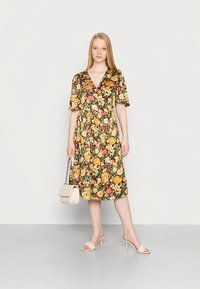 Vestido envelope floral com mangas curtas bufantes nos tons verde, amarelo e vermelho, com cintura marcada e saia até ao joelho. Acessorizado com uma pequena mala e sandálias estilo slide.