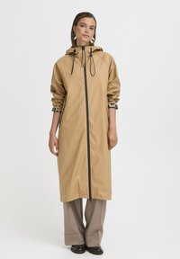 Manteau imperméable beige avec capuche, fermeture éclair à l'avant, poignets élastiques et doublure en léopard. Design long et droit avec une texture lisse.