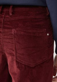 Bordeaux corduroy broek met verticale ribbelstructuur, voorzien van een achterzak en een klein metalen accent nabij de bovenkant.