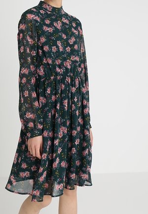 Květinové šaty z tmavě zelené látky, s dlouhými rukávy, vysokým výstřihem, staženým pasem a rozšířenou sukní s růžovými a žlutými květy.