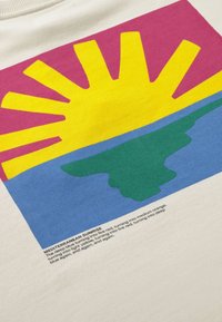 Soleil avec des rayons au-dessus d'une terre verte et d'une mer bleue dans un ciel rouge, intitulé « Lever de soleil méditerranéen », imprimé sur un tissu beige avec un texte descriptif en dessous.