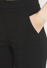 Pantalon noir à texture lisse, avec une ceinture large et des poches latérales ; des lignes épurées et une coupe ajustée mettent en valeur le design.